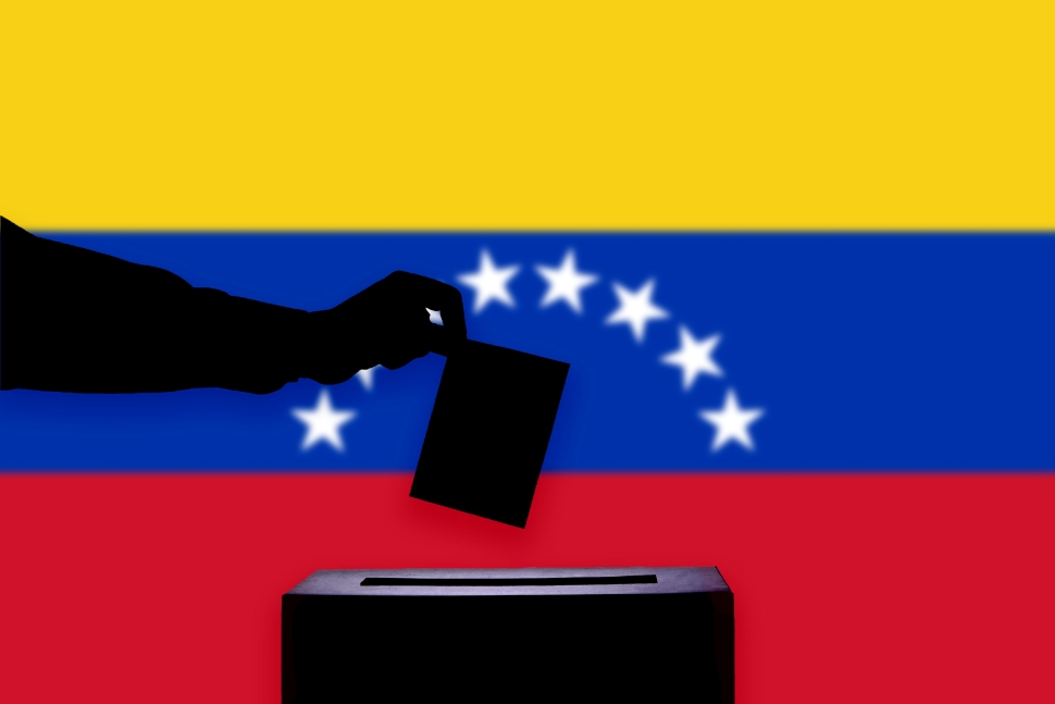 La silueta de una mano emitiendo un voto en una urna se representa frente a la bandera venezolana, que presenta franjas horizontales amarillas, azules y rojas con estrellas blancas que forman un arco en el centro. Elecciones venezolanas.