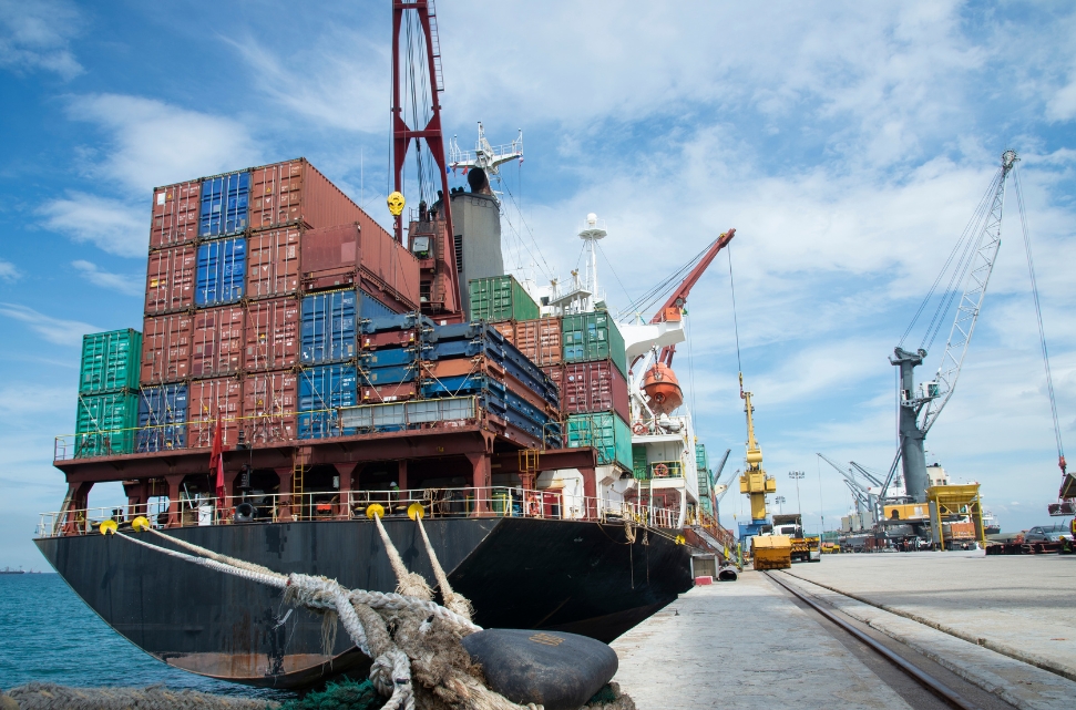 A large cargo ship docked at a port is loaded with colorful shipping containers. Cranes and machinery are visible in the background under a partly cloudy sky.